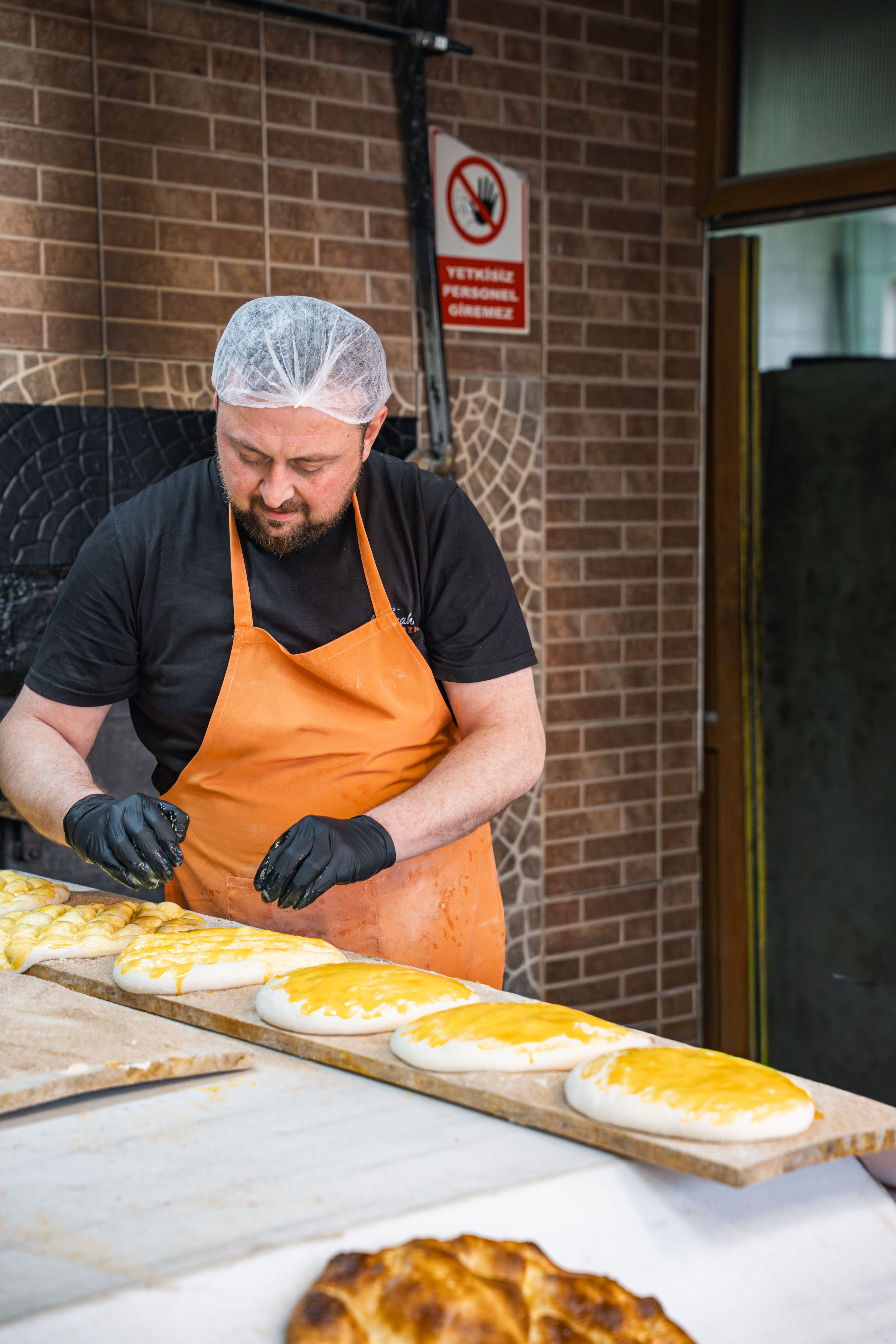 Baker wearing orange apron prepares bread in an artisanal bakery, adding a fresh coat before baking.