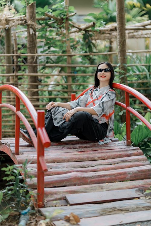 Free A stylish woman in sunglasses sits on a rustic red bridge surrounded by lush greenery. Stock Photo