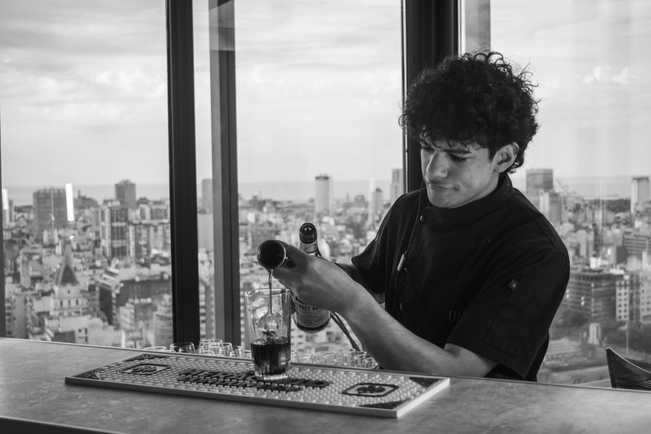 A bartender pours a drink from a bottle at a rooftop bar overlooking a sprawling cityscape.