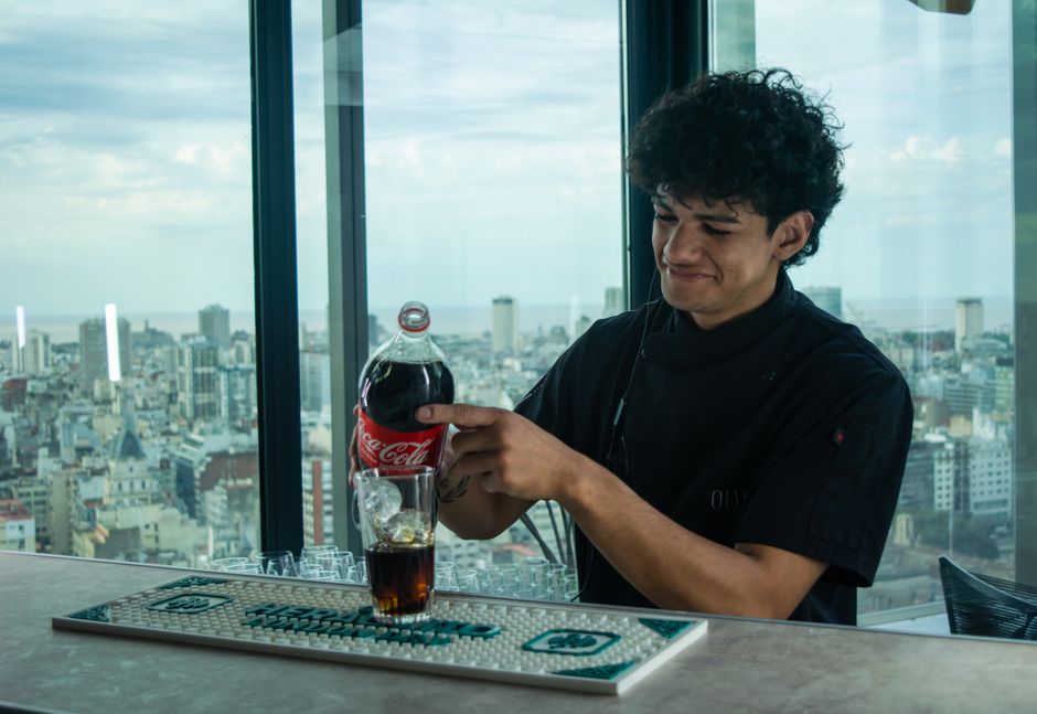 Bartender with curly hair pouring Coca-Cola in a high-rise bar with city skyline view.