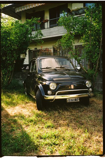 A classic black Fiat 500 parked in a sunlit garden outside a European home.