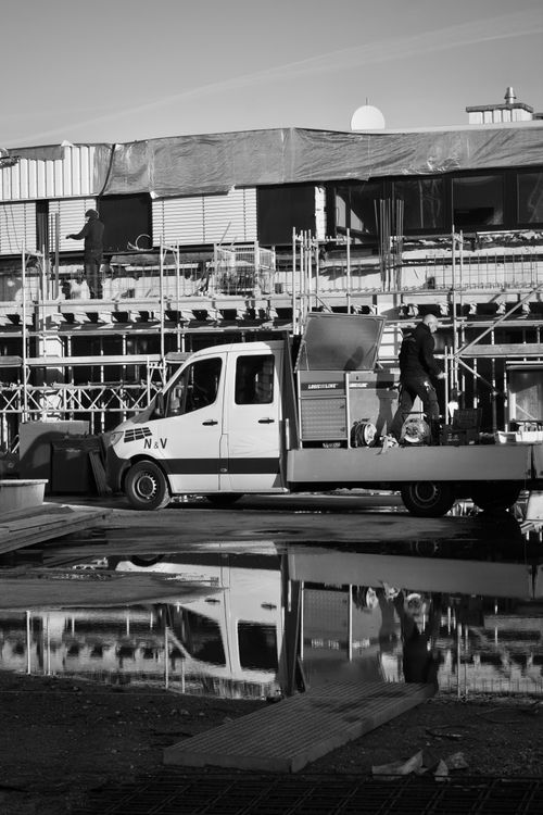 Free Monochrome image of a construction site with workers and vehicles reflecting in water. Stock Photo