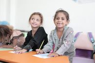 Two young children smiling while engaging in a classroom activity with pencils and paper.
