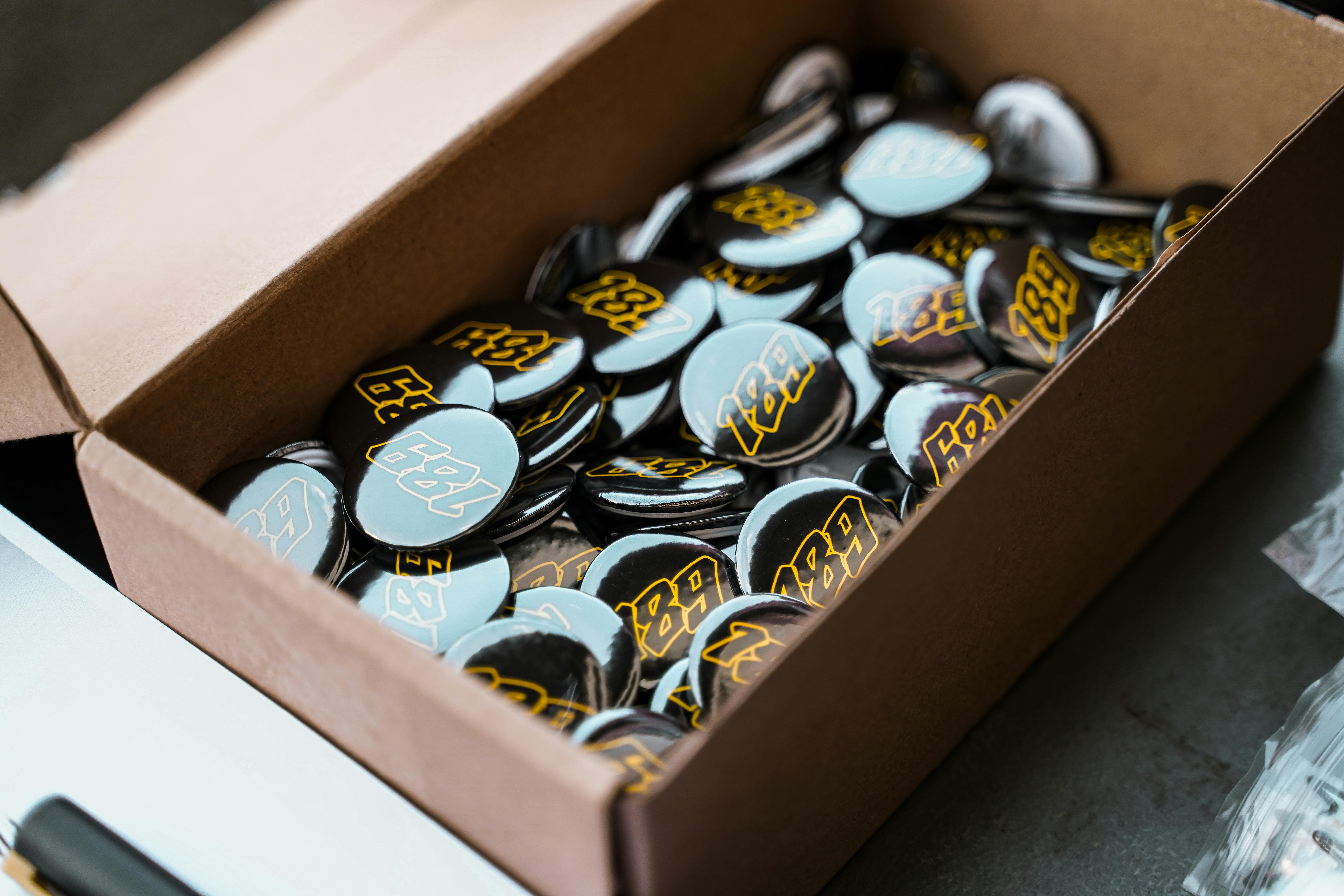 A cardboard box filled with shiny, circular pins featuring the number 182 in bold text.