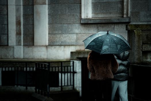 A couple shares an umbrella on a rainy day in front of an urban building. Moody and intimate city scene.