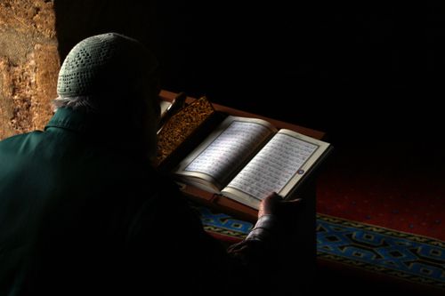 Free An elderly man reading the Quran in a peaceful, dimly lit room, focused on spiritual connection. Stock Photo
