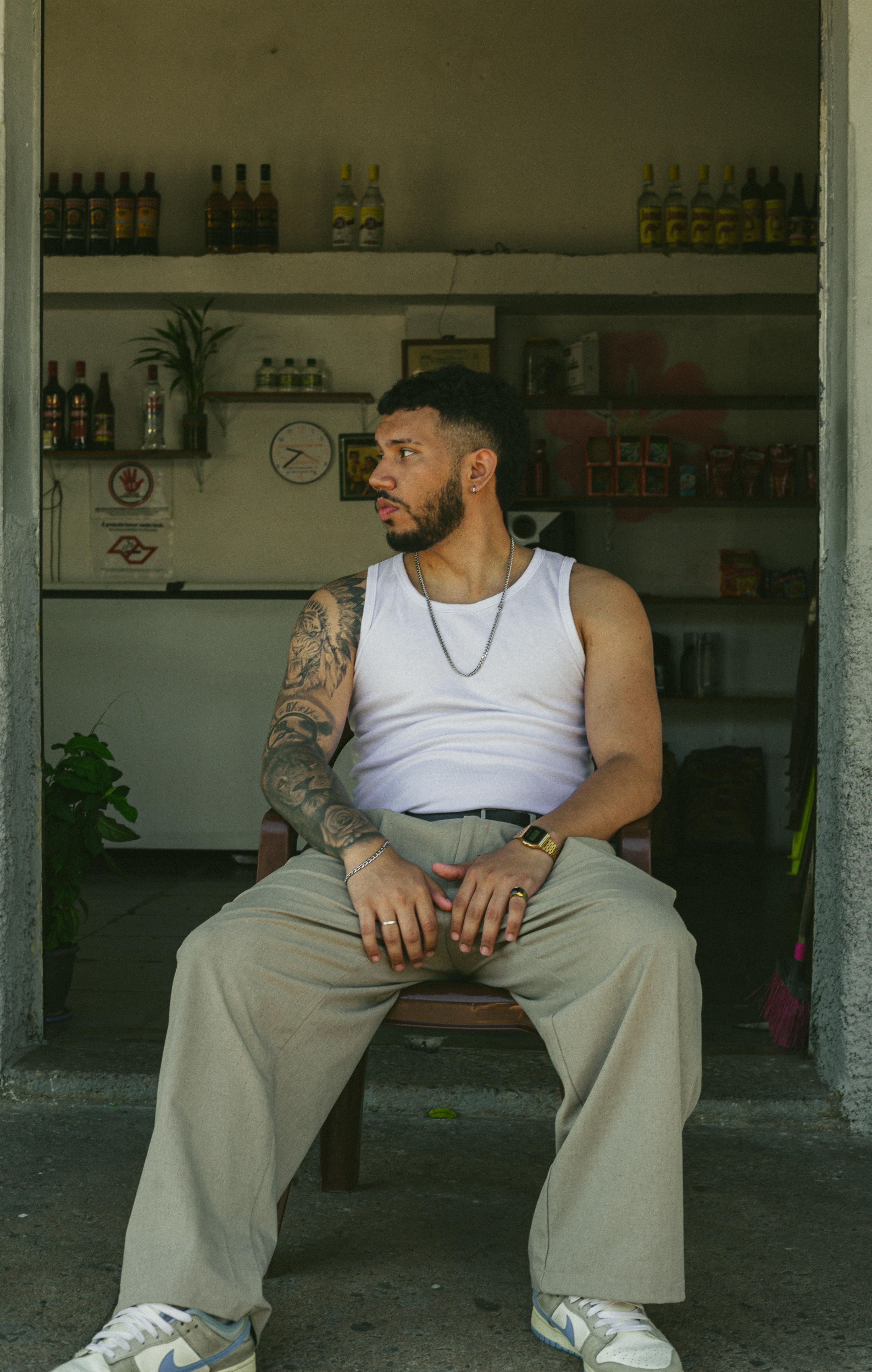 Young man with tattoos in casual streetwear sits outside a rustic shop in São Paulo.