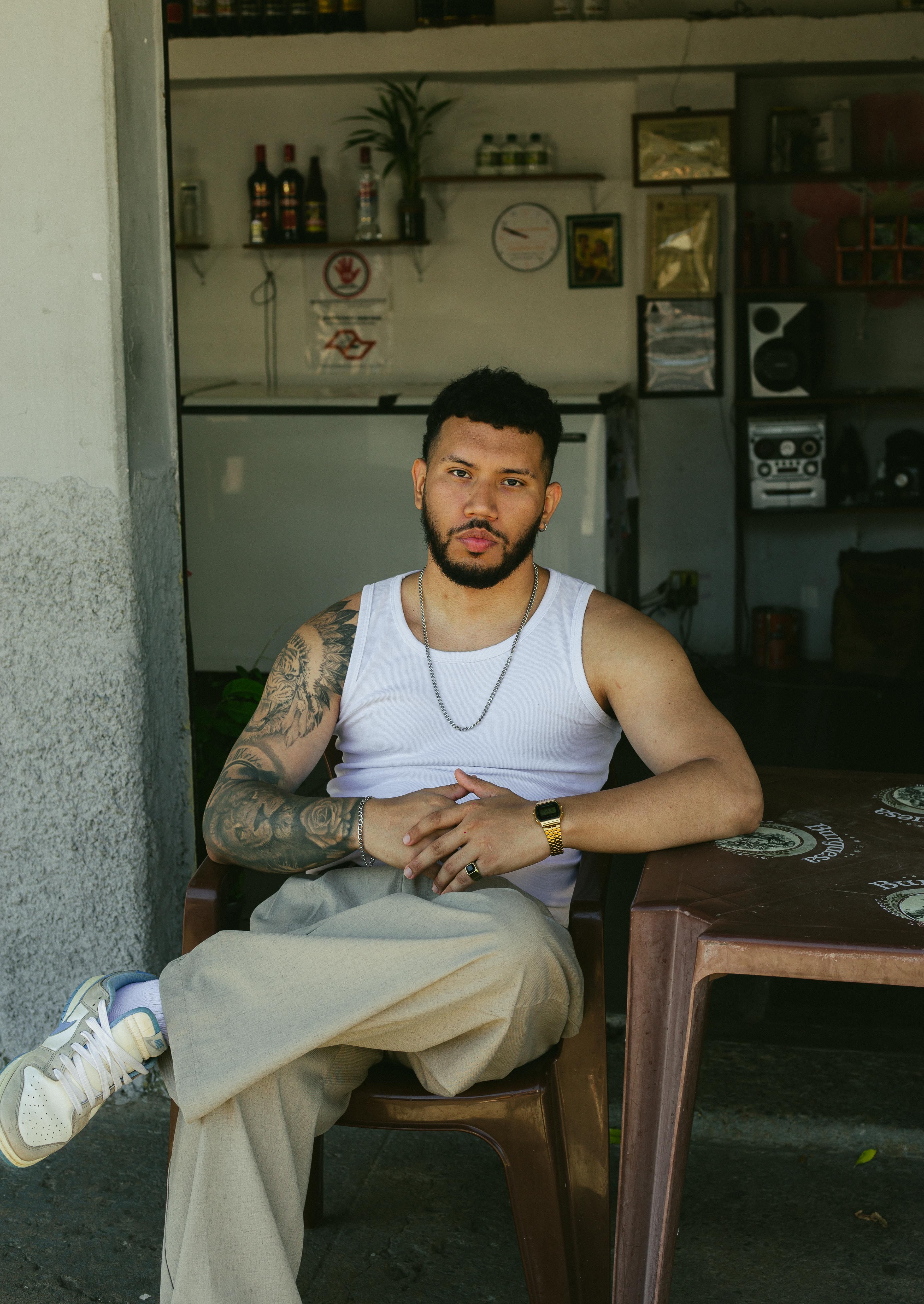 A fashionable man with tattoos relaxes in a Brazilian street bar wearing a white singlet and casual style.