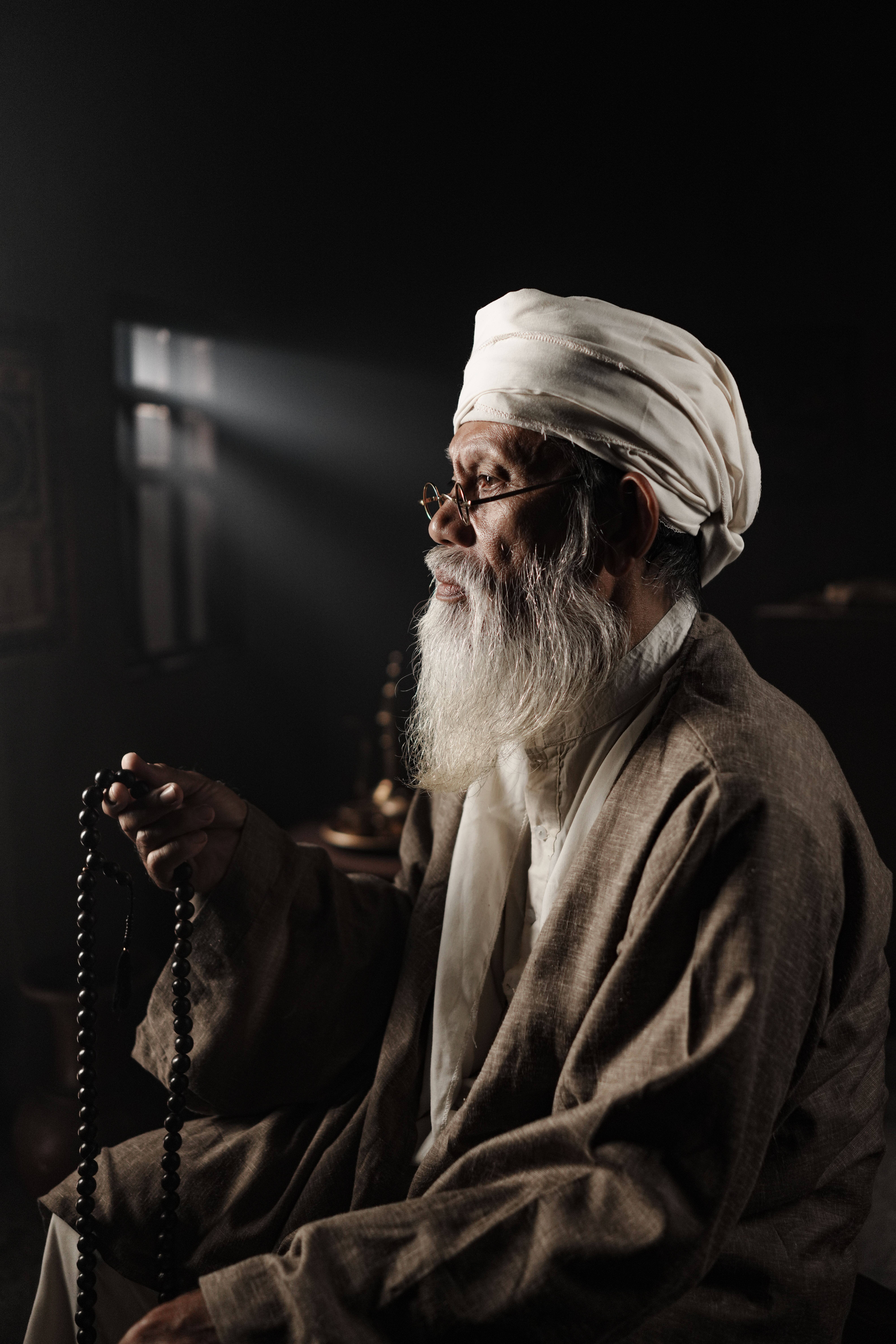An elderly man in traditional clothing holds prayer beads in a dimly lit room, exuding spirituality.