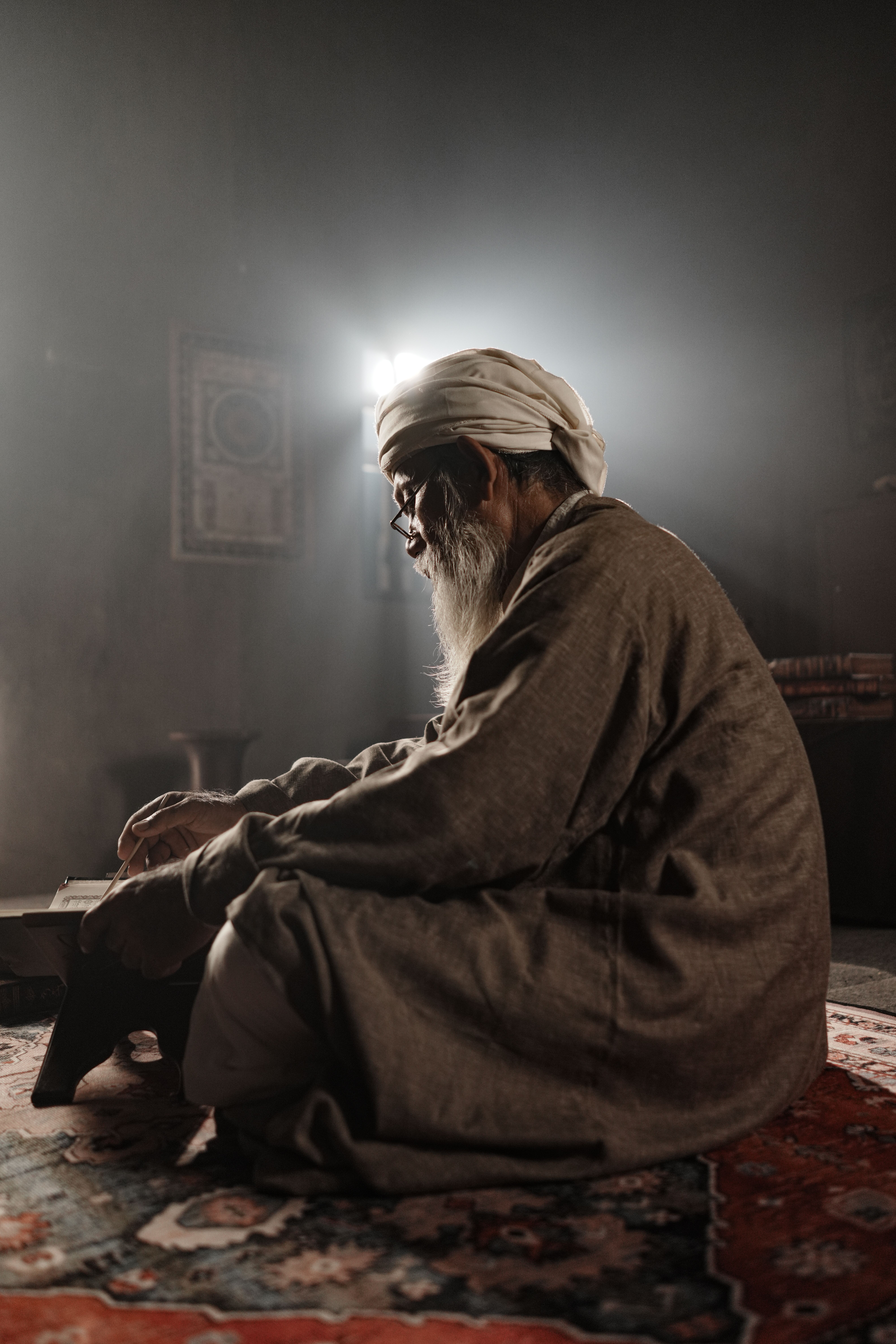 Elderly man in traditional attire, seated on ornate carpet, praying in dimly lit room.