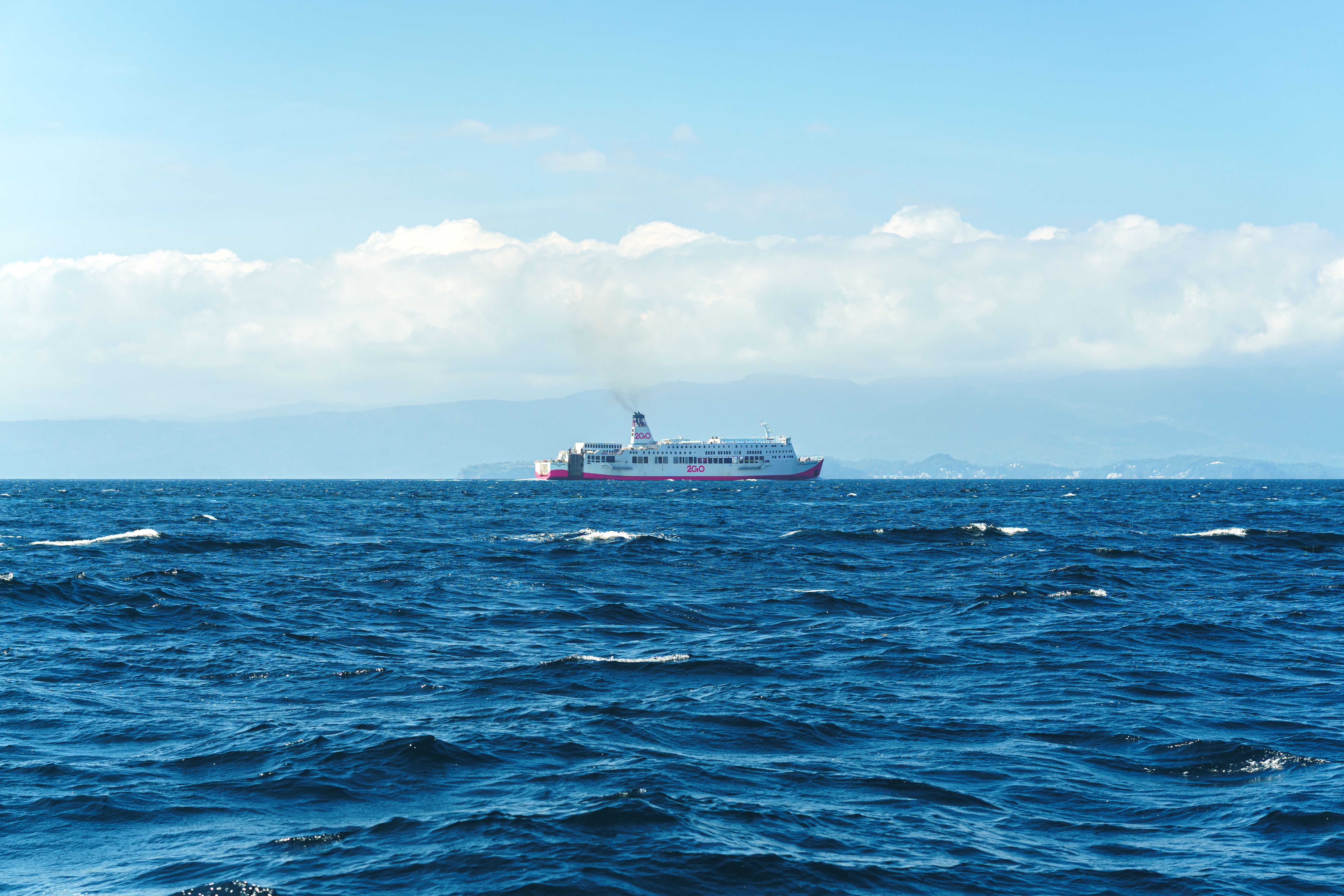 A lone ferry travels across the vast, blue ocean under a bright sky, emitting a trail of smoke.