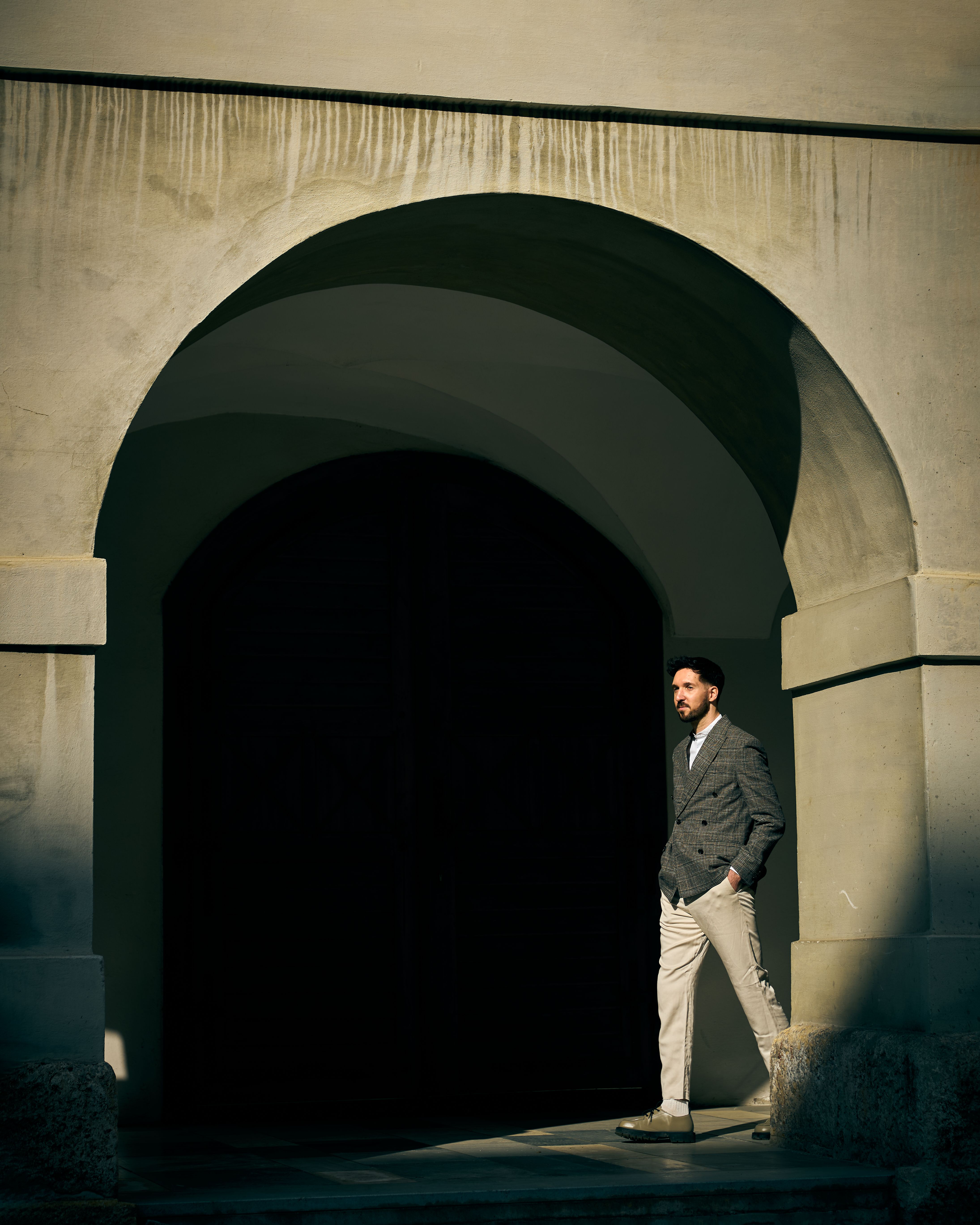 A fashionable man in a blazer walking through a sunlit archway in Graz, Austria.