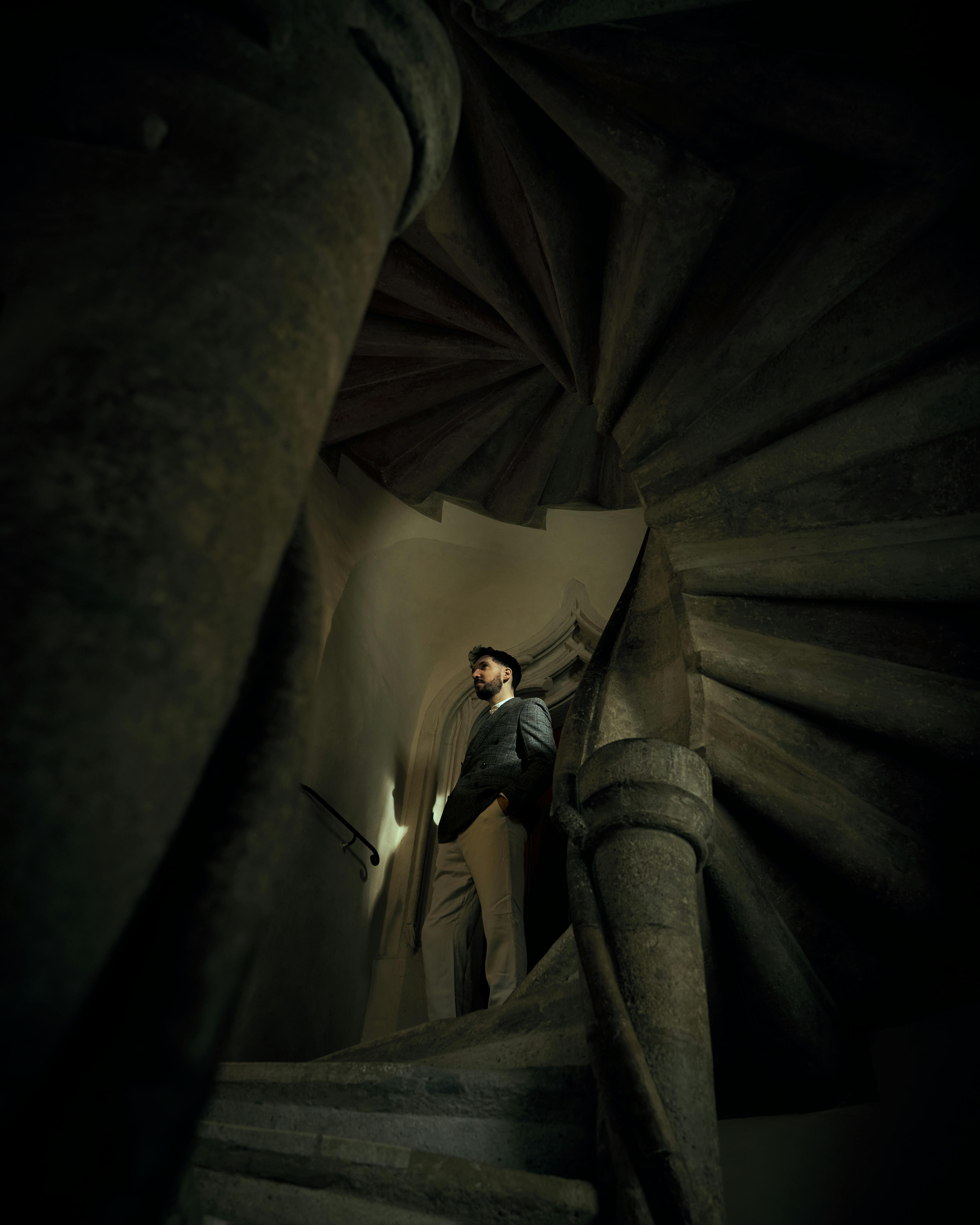 grátis Um homem está em pé em uma antiga escadaria em espiral, criando uma composição arquitetônica melancólica em Graz, Áustria. Foto profissional