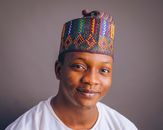 A cheerful young man wearing a colorful Nigerian cap and white attire, smiling softly.