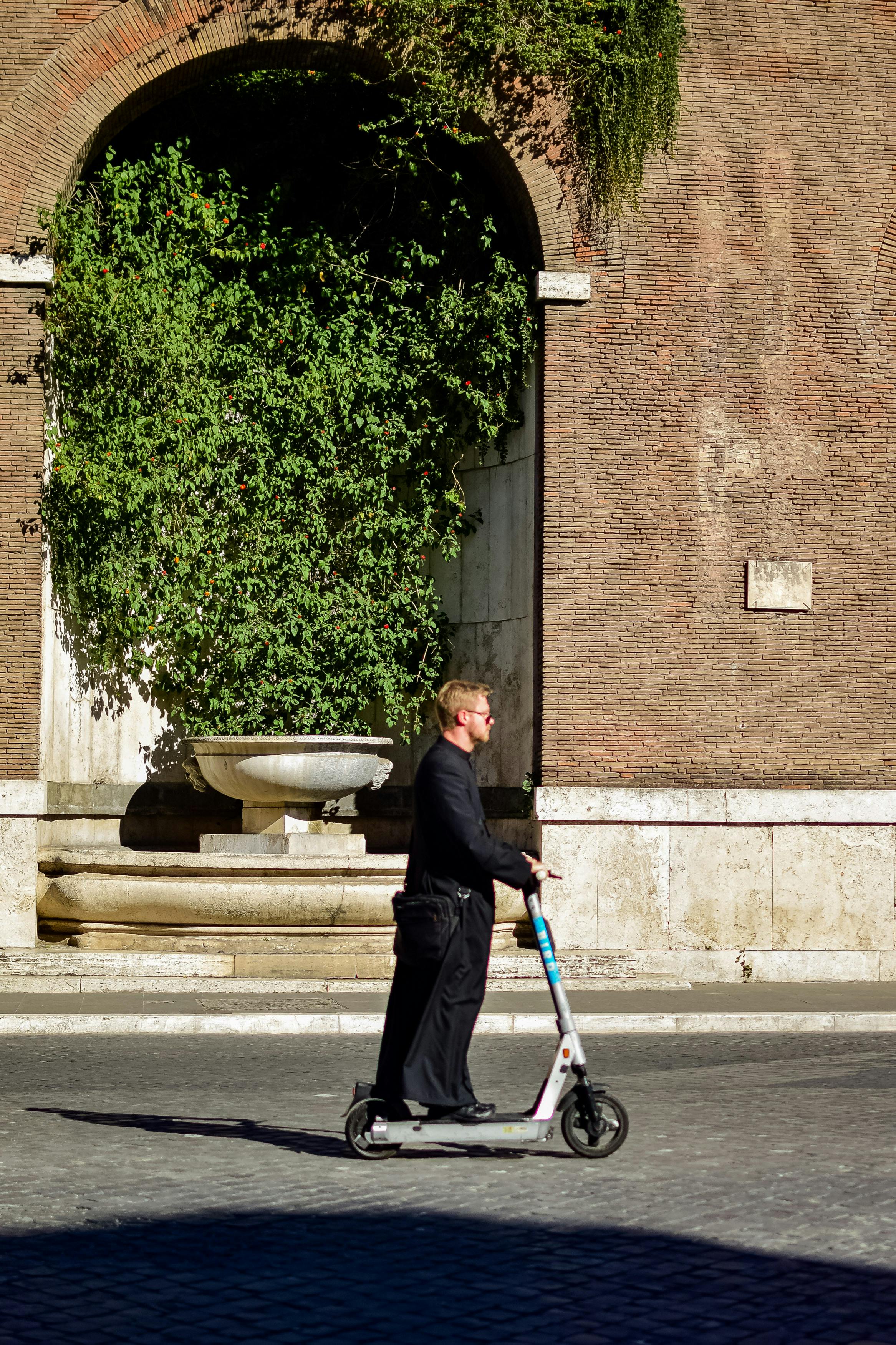 A man rides a scooter past a historic Roman wall and fountain.