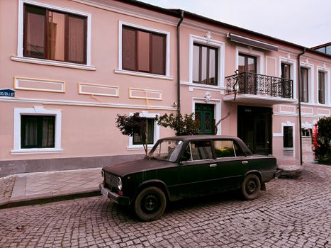 Old black car parked on a cobblestone street in front of a pastel building with balcony.