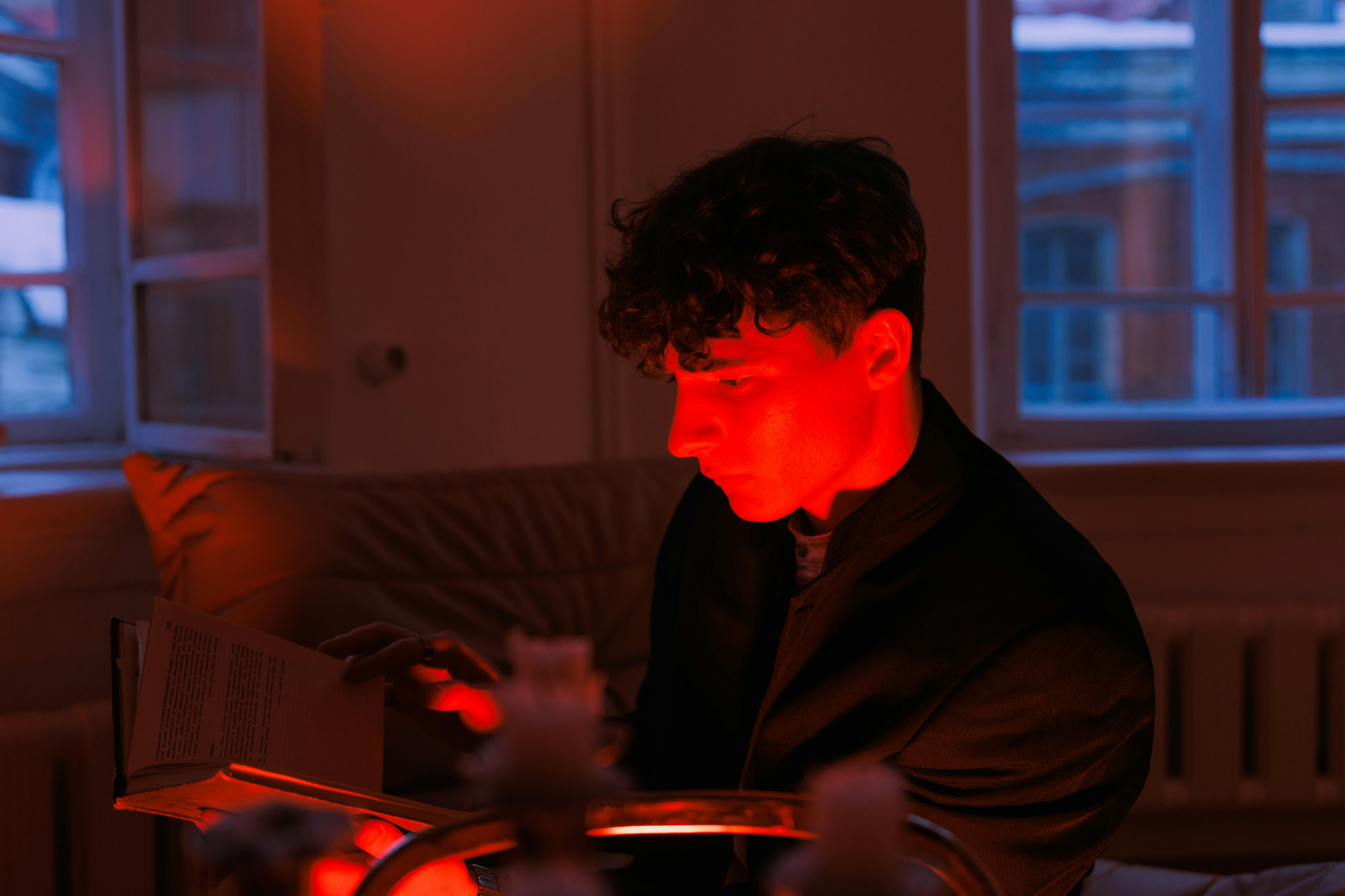Young man immersed in reading indoors with dramatic red lighting creating a moody atmosphere.