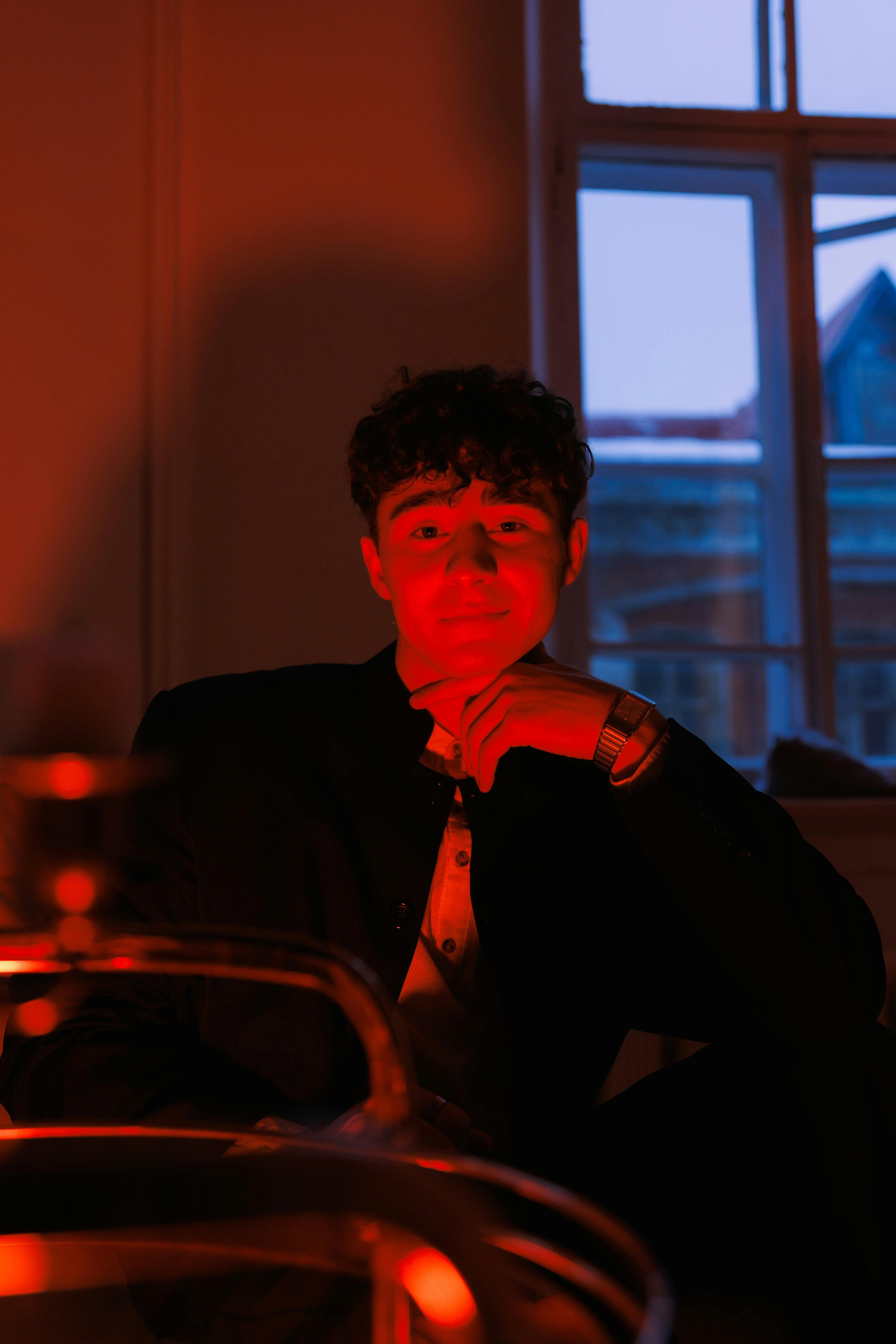gratis Portret van een jonge man, verlicht door rood licht, wat een sfeervolle ambiance creëert binnenshuis, met uitzicht naar buiten door het raam. Stockfoto