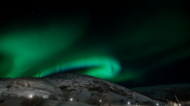 Capture of the Northern Lights displaying vibrant green hues over a snow-covered hill at night.