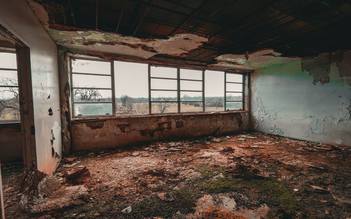 A dilapidated room showing crumbling walls, broken windows, and decayed floor with a distant view of trees.