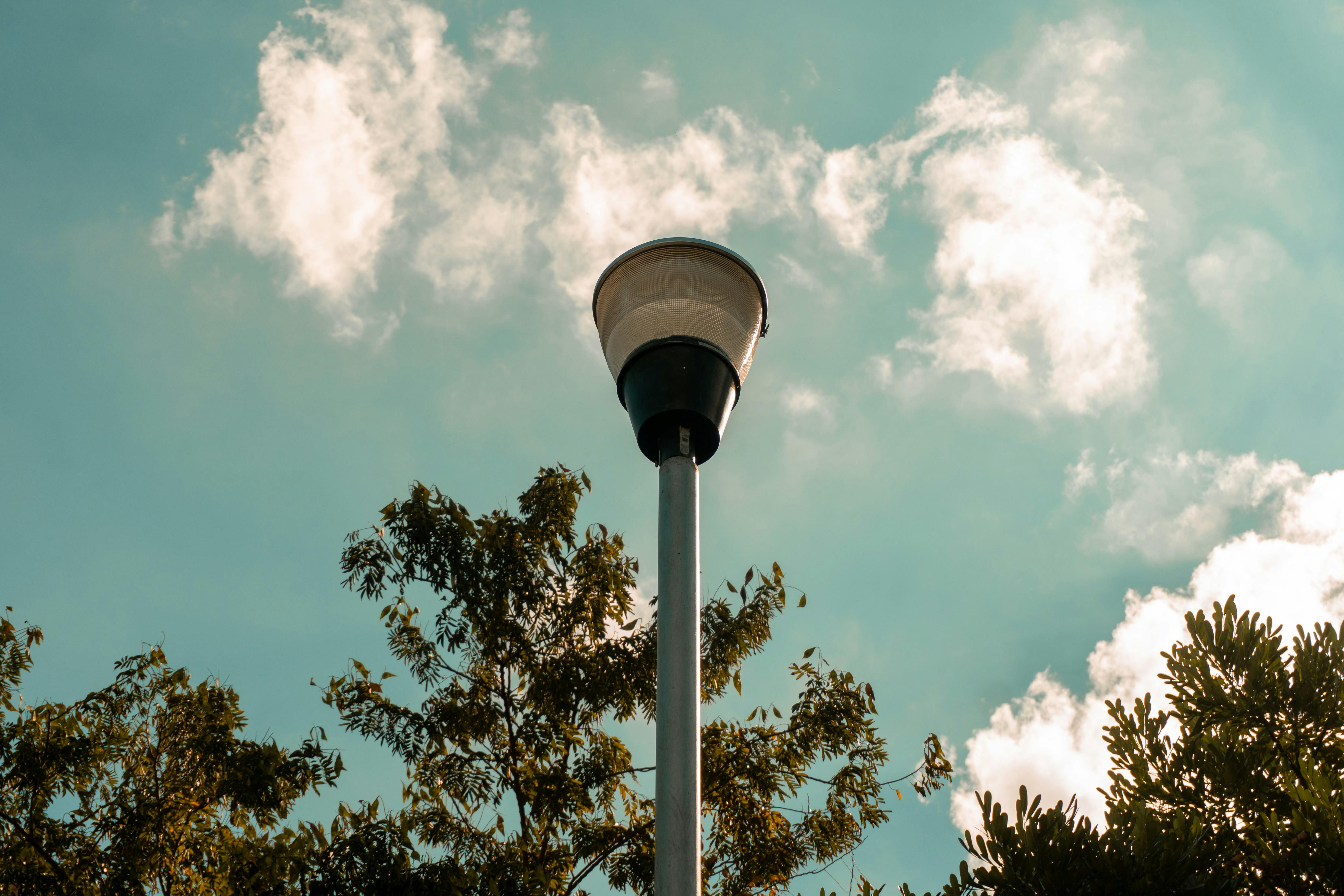 Zadarmo Pouličná lampa sa týči vysoko na pozadí oblačnej modrej oblohy, obklopená listami stromov. Fotka z fotobanky