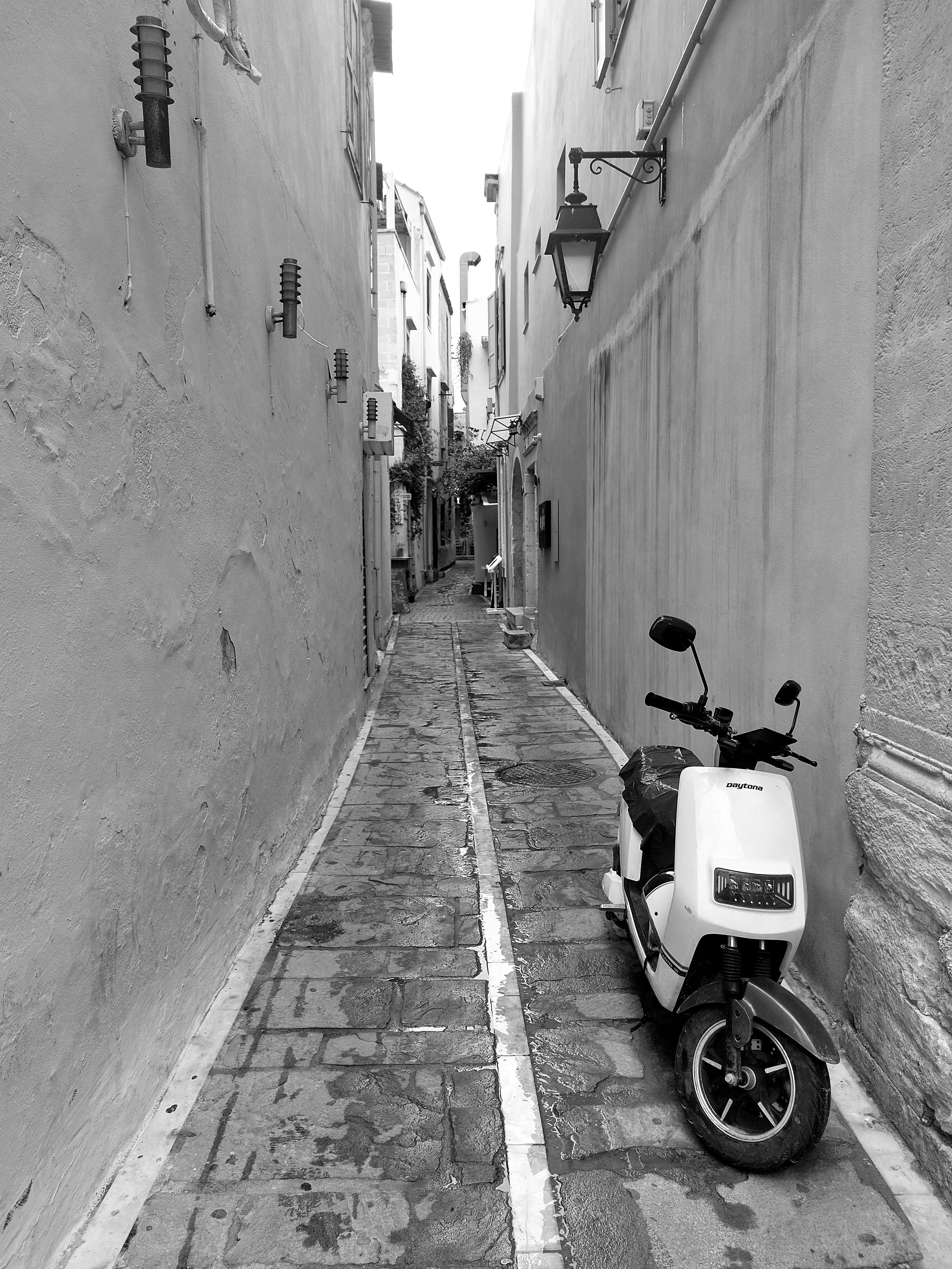 Bezpłatne Czarno-białe zdjęcie wąskiej, brukowanej uliczki z zaparkowanym skuterem, przywodzące na myśl europejski urok. Zdjęcie z galerii