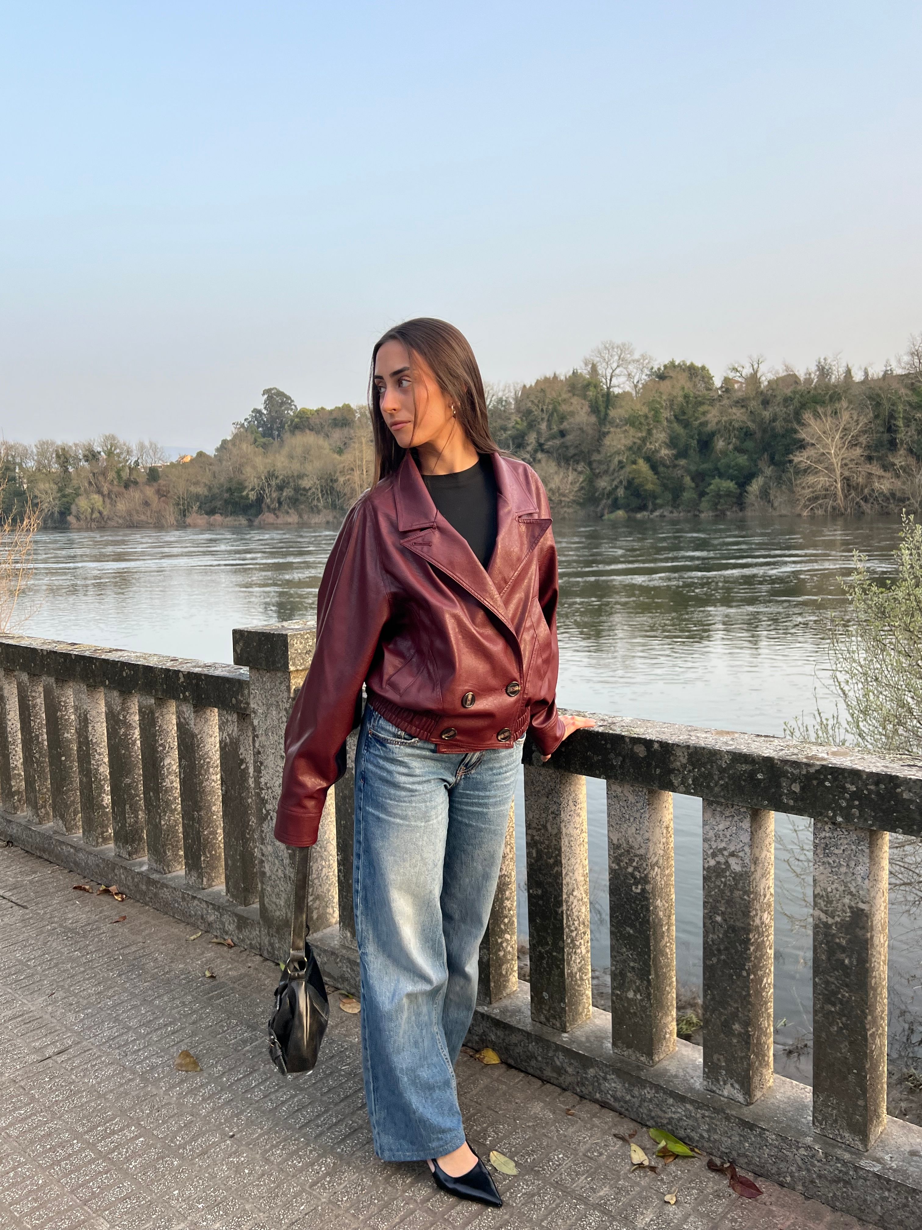 Young woman in a red leather jacket stands by a river with lush greenery.