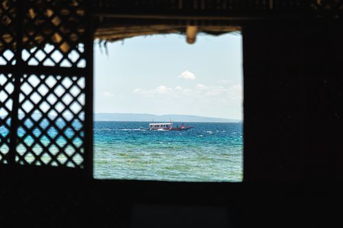 Free A serene view of the sea with a boat framed by a lattice window from a shaded room. Stock Photo