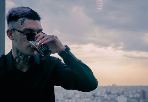 Free Tattooed man with sunglasses drinks soda in urban setting at sunset. Stock Photo