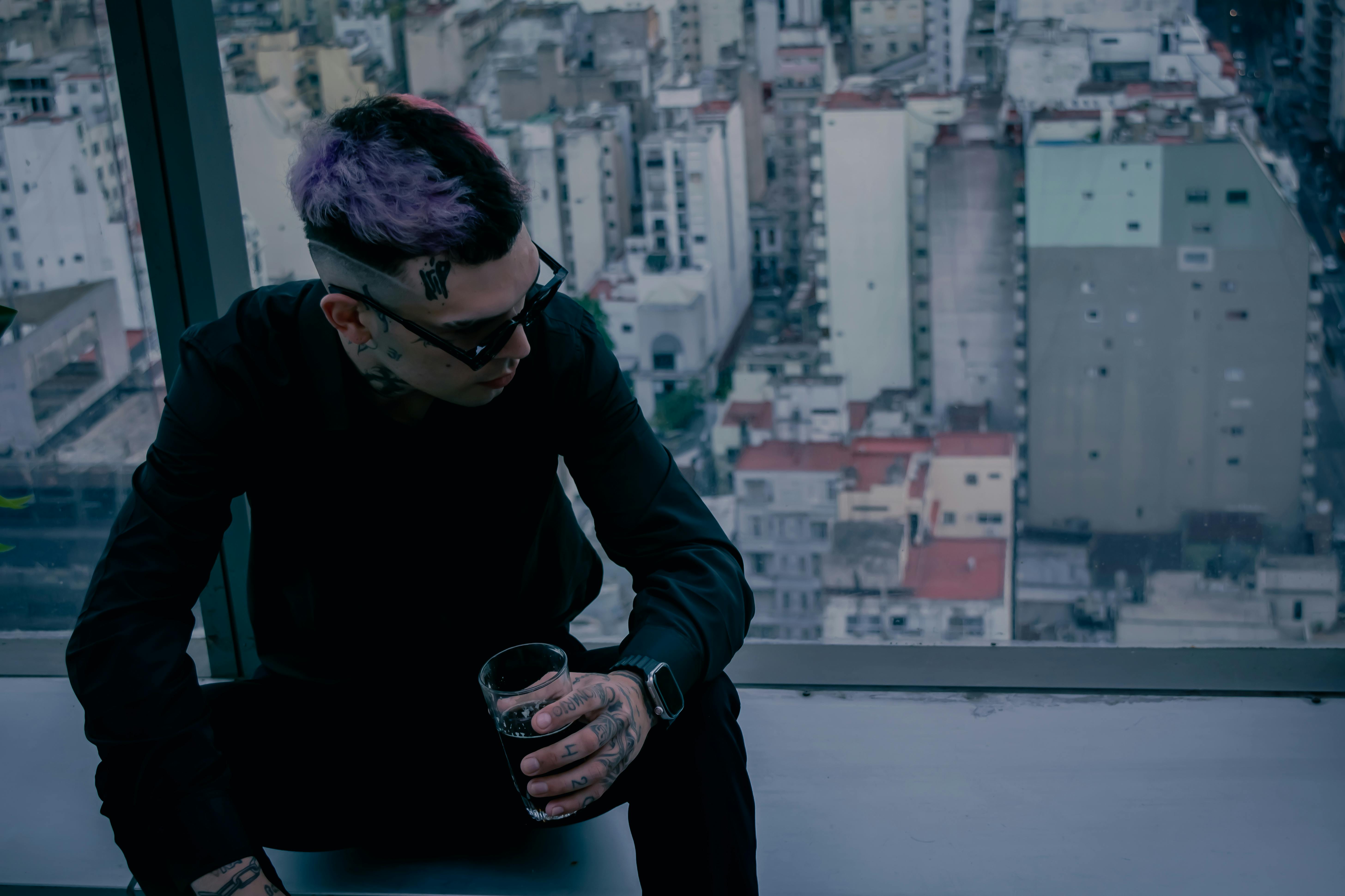 Gratis Un uomo elegante e tatuato, vestito di nero, è seduto vicino a una finestra, da cui si gode una vista panoramica sulla città. Foto a disposizione