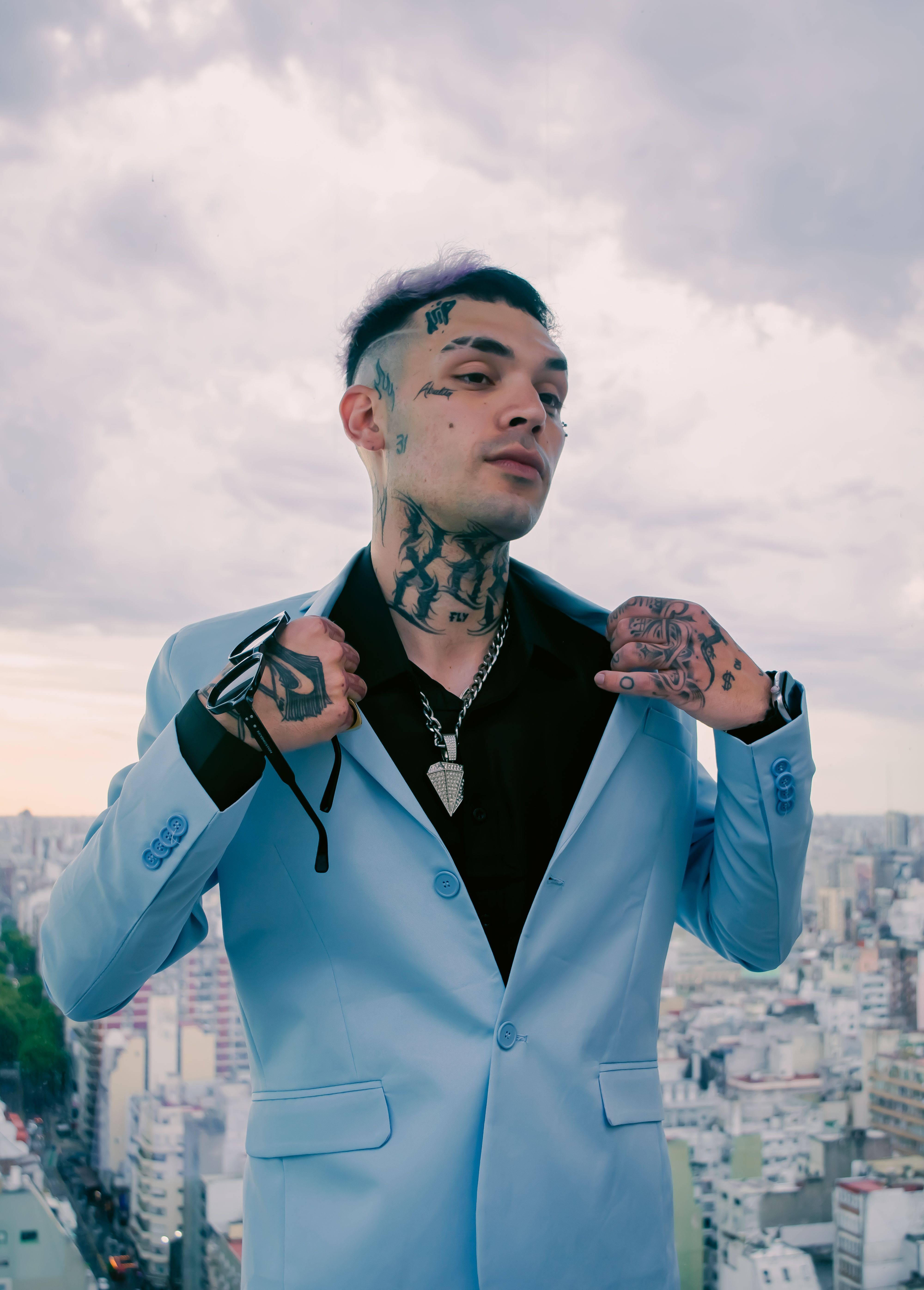 Young tattooed man in a blue blazer poses confidently on a city rooftop, showcasing urban style and edgy fashion.