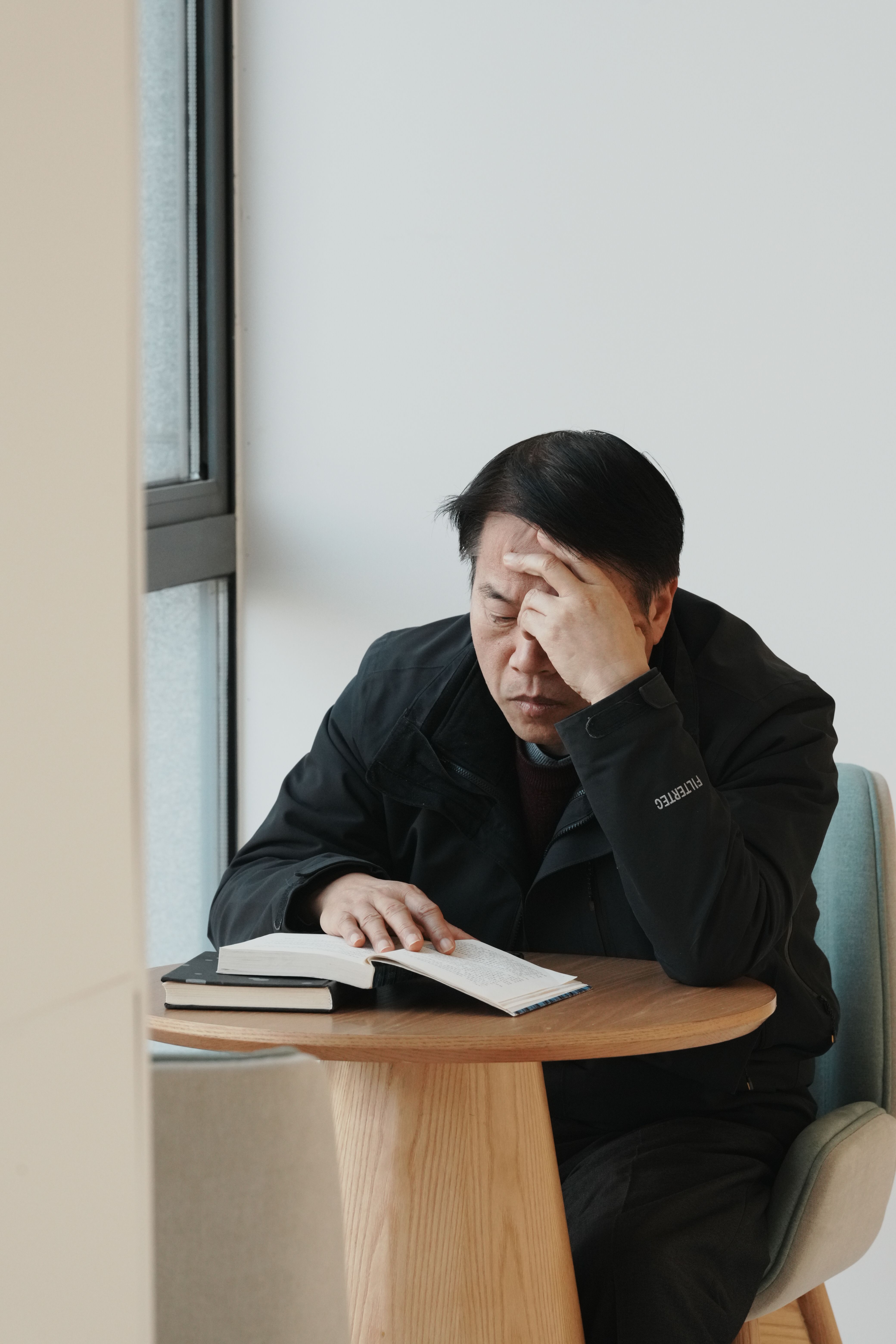 An adult man in deep thought holding his head while reading a book at a table indoors.