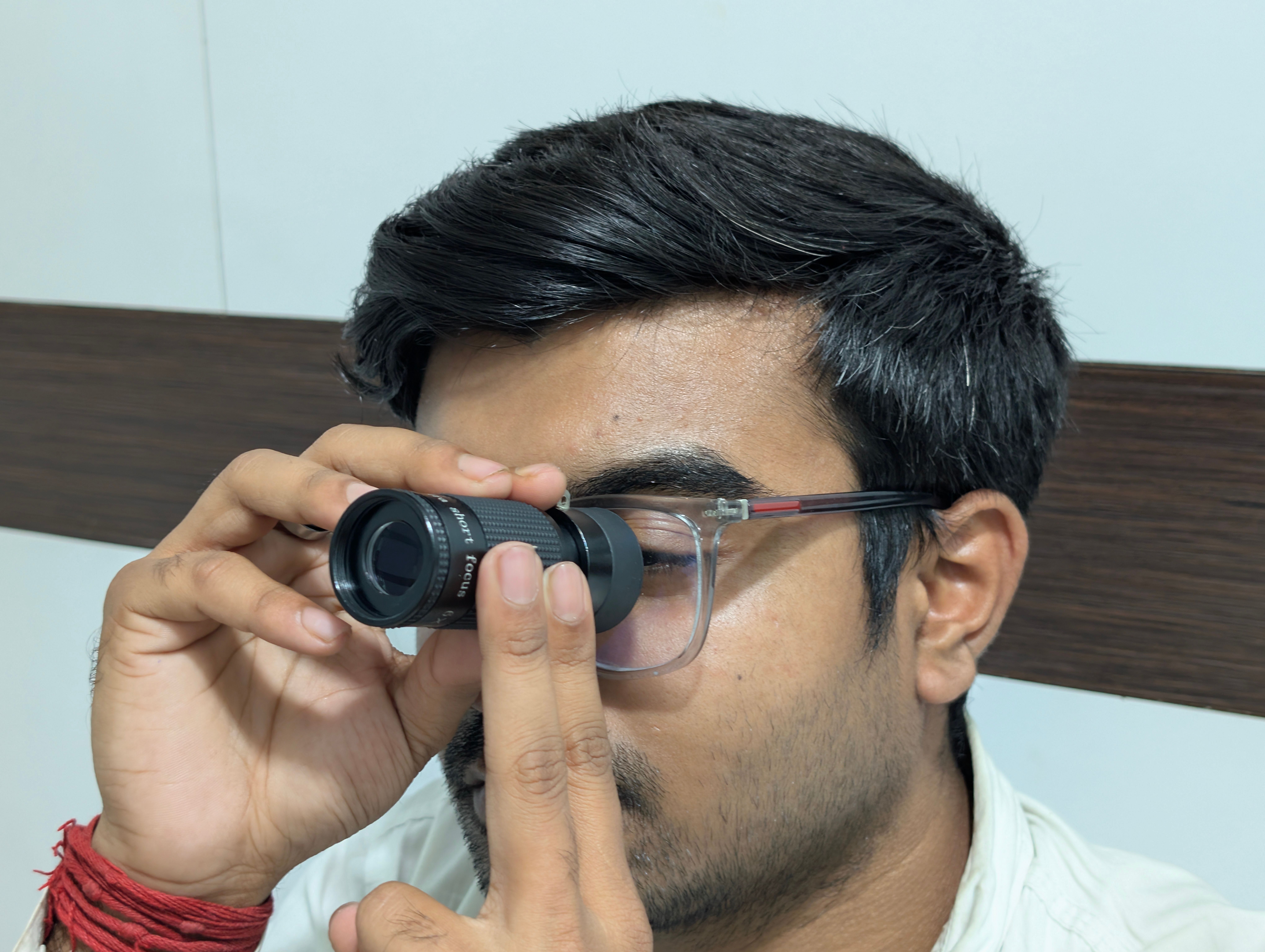 A man examines an object using a handheld telescope indoors, focusing closely.