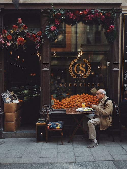 Man in Brown Jacket Sitting on Chair in Front of Fruit Stand