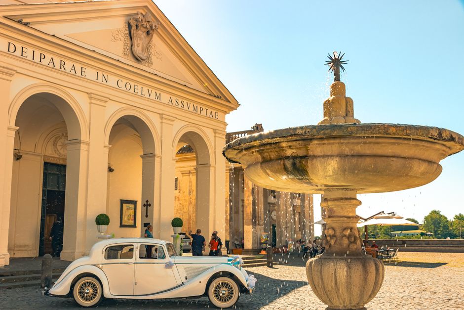 A classic car parked in front of a historic neoclassical building with a fountain.