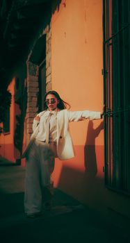 Confident woman in a white suit posing outdoors against a sunlit orange wall.