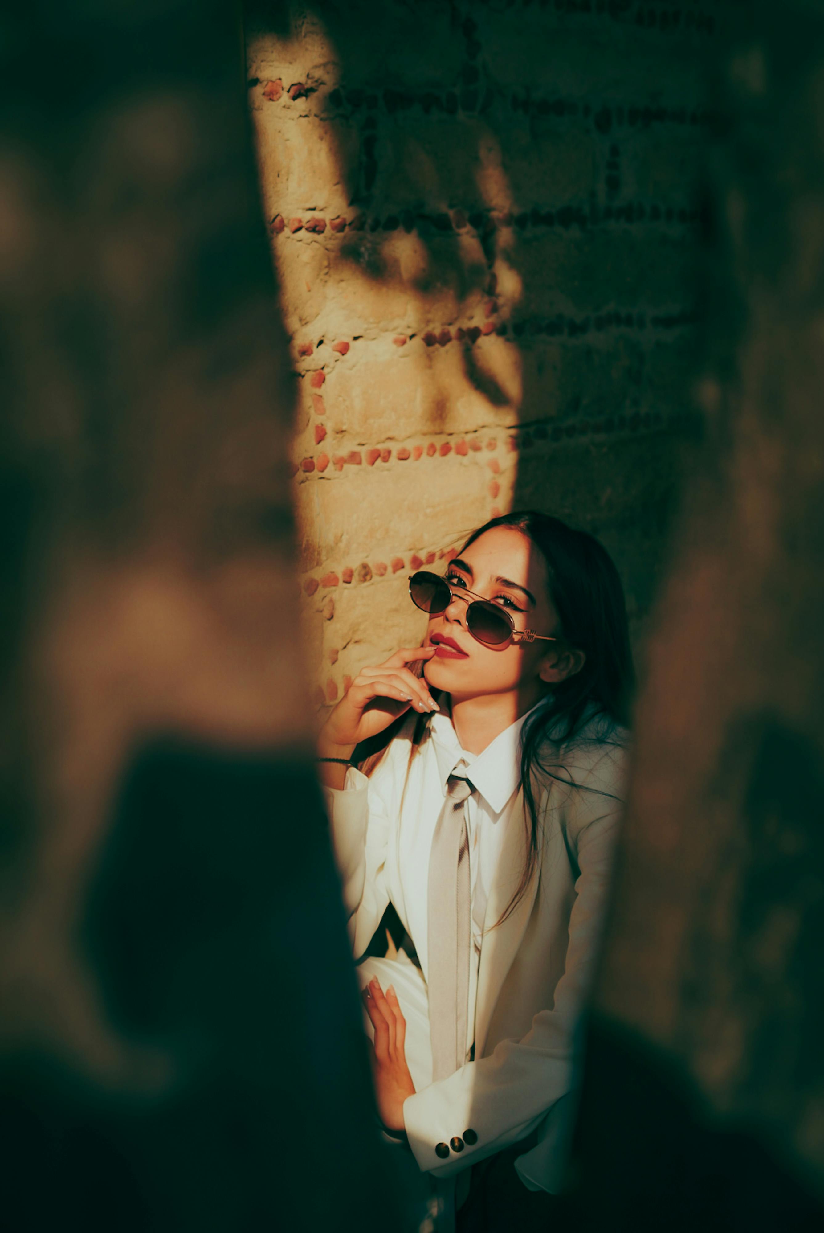 無料 白いスーツとサングラスをかけた女性が、ドラマチックな光と影のある素朴な壁を背景にポーズをとっています。 写真素材