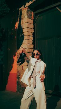 Trendy woman in a white suit poses stylishly outdoors with sunglasses and heels.