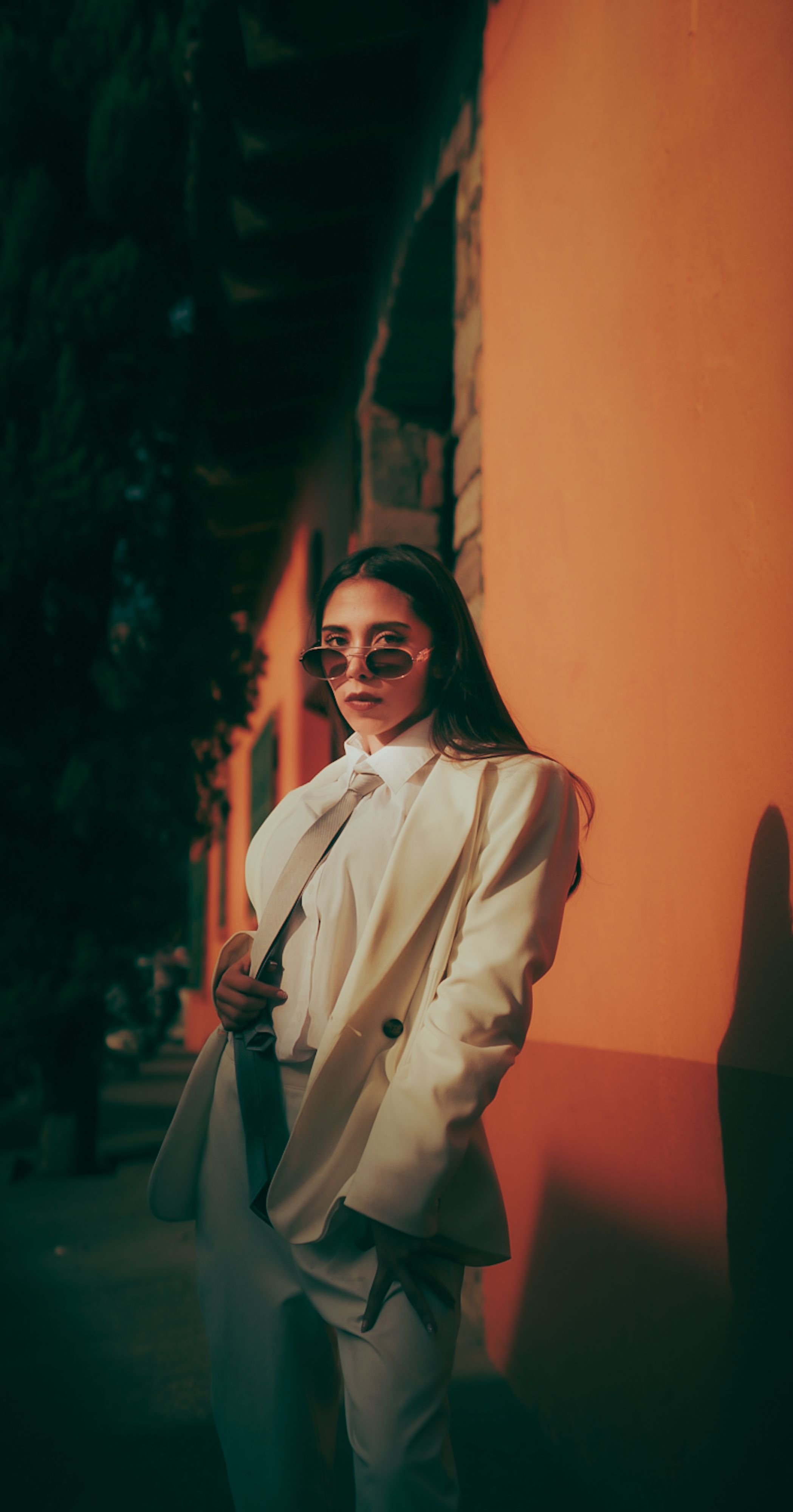 Mulher fashionista em um terno branco e óculos de sol posando contra um fundo laranja ao ar livre.