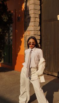 Chic woman in a white suit and sunglasses striking a pose on a sunny city street.