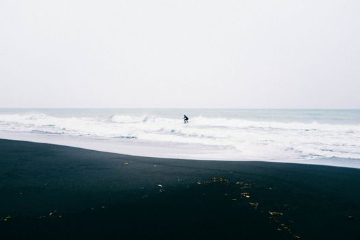 Lone surfer catching waves on a serene black sand beach with gentle ocean waves.
