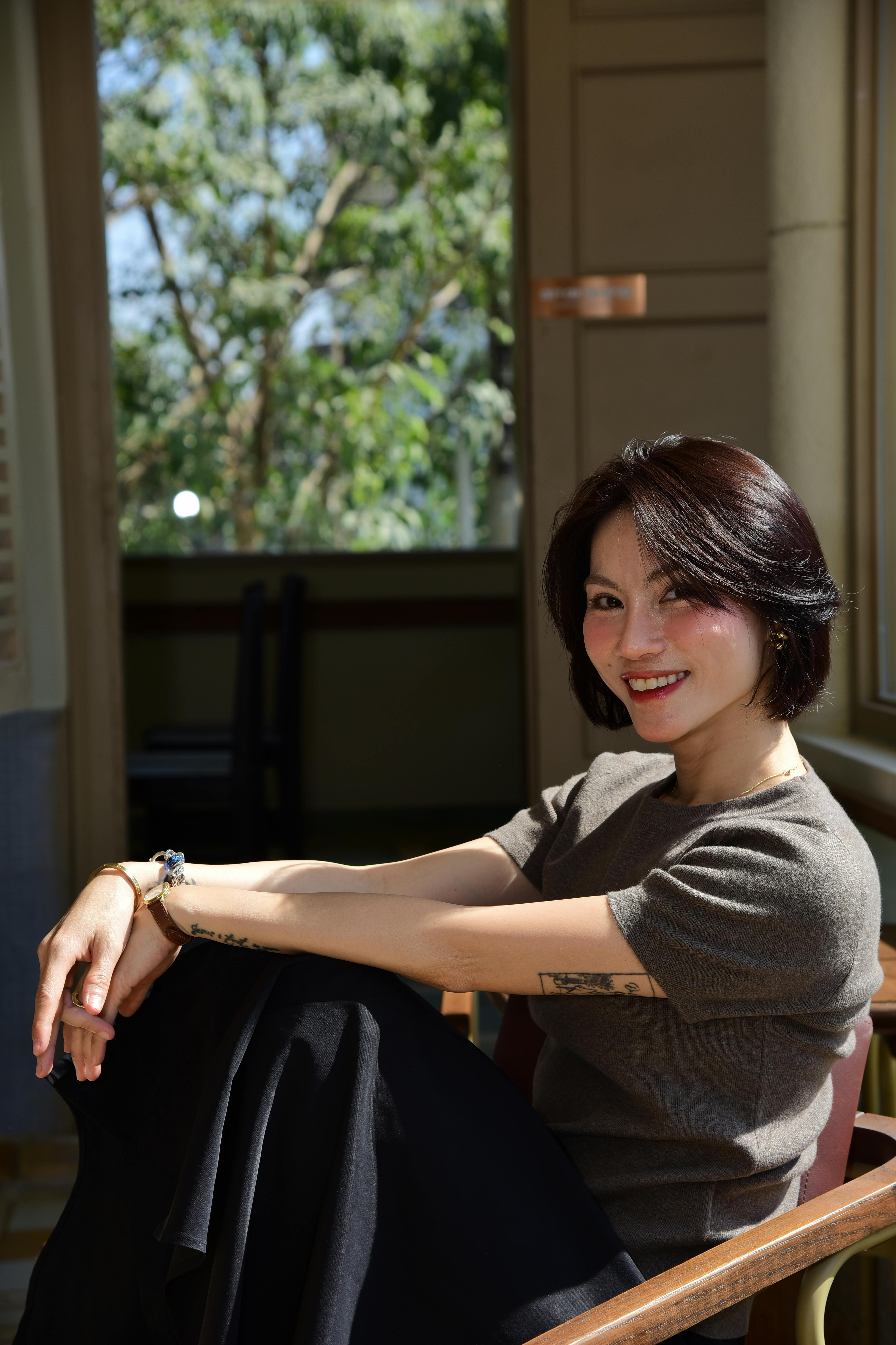 A cheerful woman sits indoors, relaxed and smiling, with sunlight streaming in.