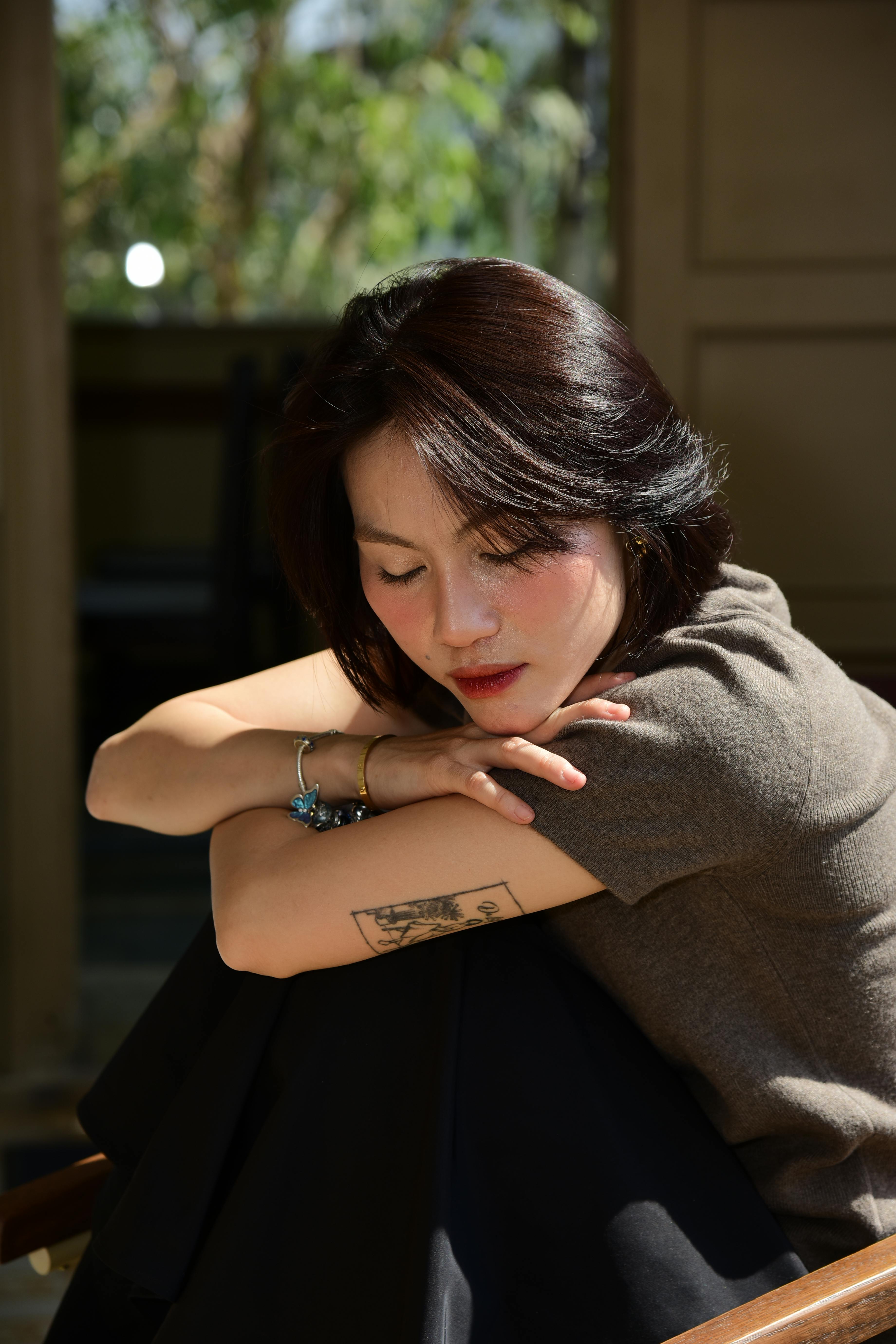 A woman sitting indoors, leaning on her arms with eyes closed, bathed in soft natural light.