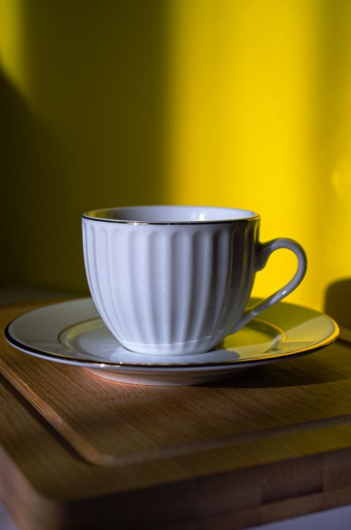 Free A stylish porcelain teacup with saucer sits on a wooden surface against a bright yellow background. Stock Photo