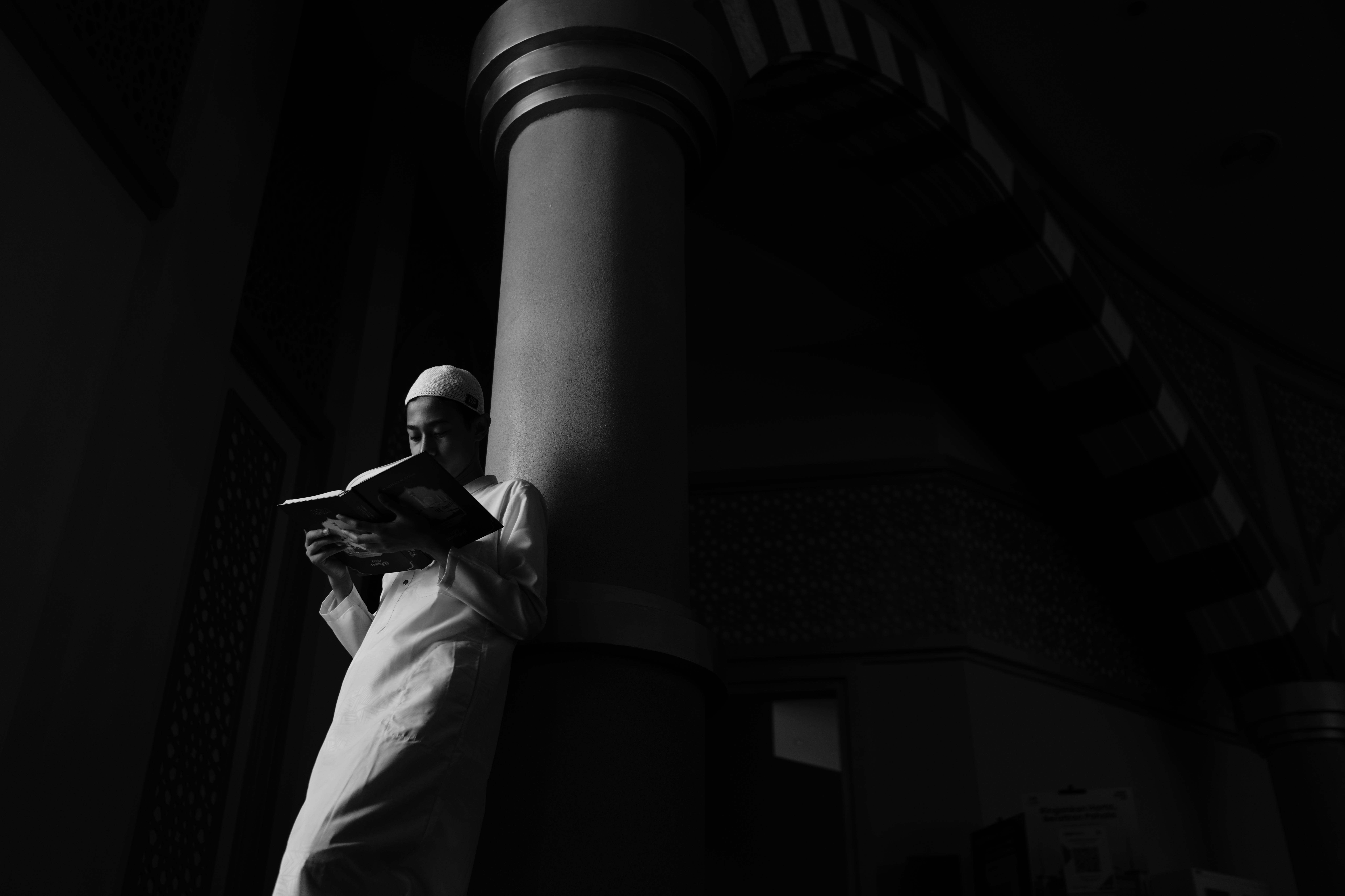 Ücretsiz Loş bir camide, derin gölgelerin yarattığı tefekkür dolu atmosferde kitap okuyan genç bir adam. Stok Fotoğraflar