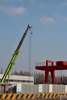 A green construction crane and red crane arm in a fenced industrial area under a blue sky.
