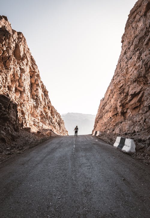 Man Walking on Asphalt Road Between Huge Rock Formation During Daytime