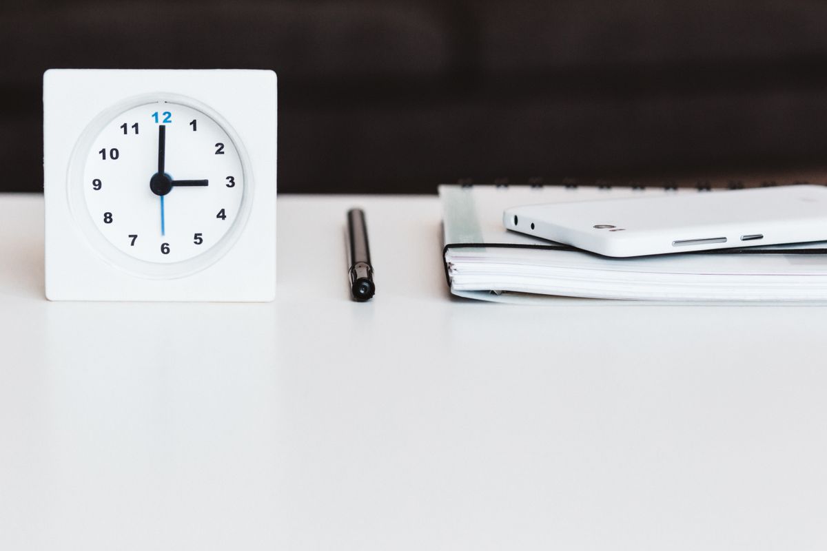 A white desk clock at twelve o'clock beside a pen, notebook, and phone on a clean desk