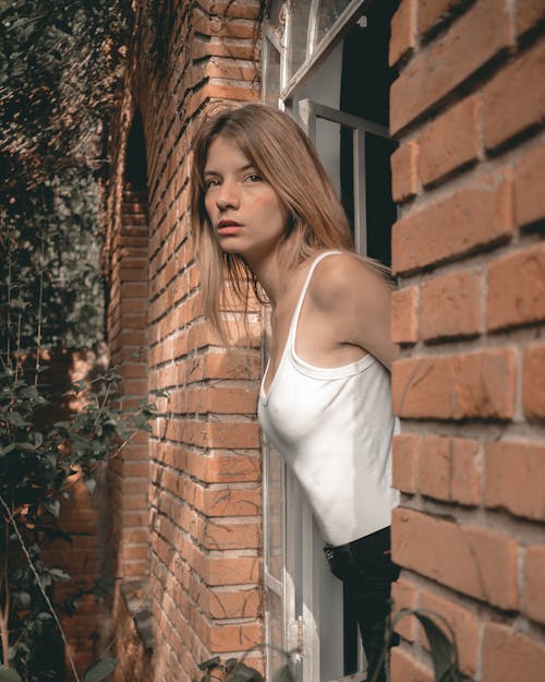 Woman in White Tank Top and Black Pants Standing Beside Brown Brick Wall