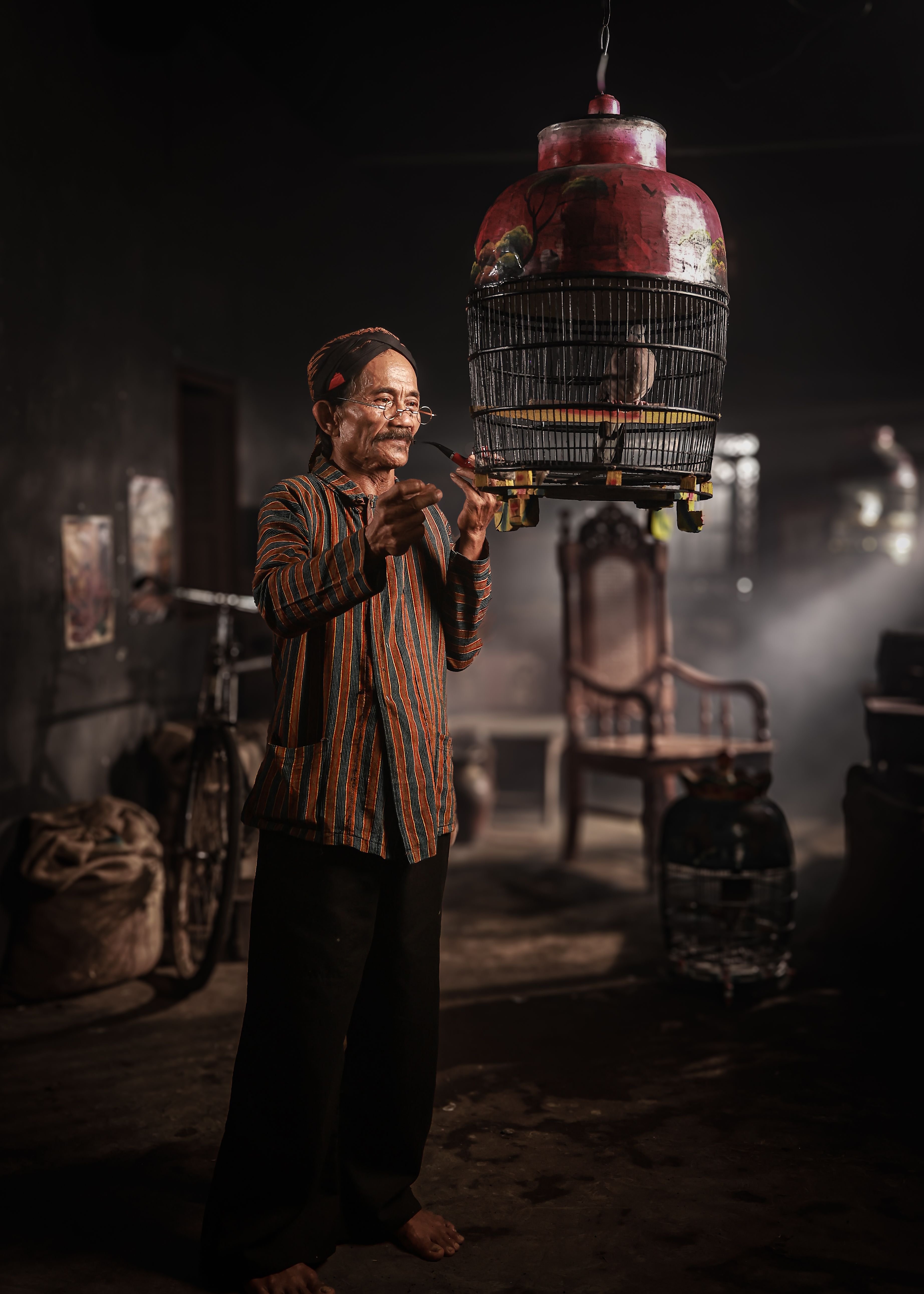 Elderly man engaging with a birdcage in a dimly lit room in Jakarta, Indonesia.